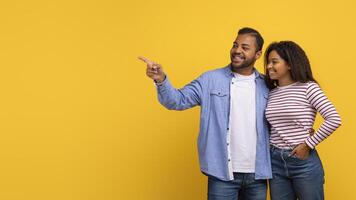 African American couple pointing at an unseen object while standing against a vibrant yellow background. They both appear engaged and focused on whatever they are indicating, copy space photo