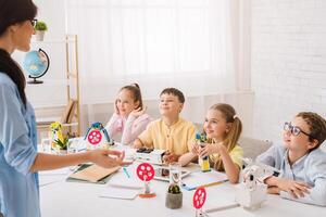 A young teacher instructs a group of elementary school students during a STEM activity. The students are sitting around a table, listening attentively to the teachers explanation. photo