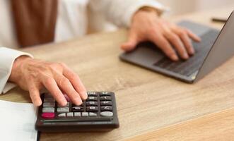Closeup Of Elderly Man's Hands Calculating Finances Using Calculator And Laptop Sitting In Office. Cropped photo