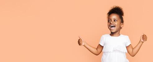 African American little girl in a white T-shirt smiling and giving a thumbs-up with both hands. She has brown curly hair, and her eyes are sparkling with joy. The background is a plain, peach color. photo