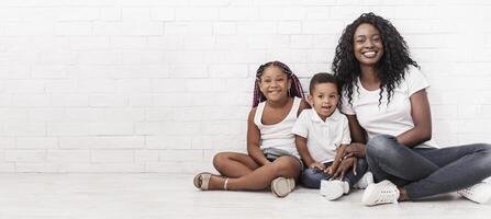 Black millennial mom sitting on floor with daughter and son and smiling at camera over white wall background. photo