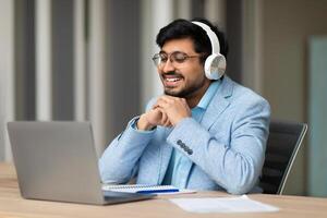 Indian man wearing headphones and glasses sits at a desk and smiles while looking at a laptop screen. He is likely participating in a call with someone else. A notebook and pen are on the desk photo