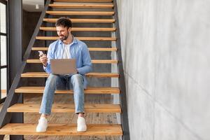 A man in a blue shirt sits on wooden stairs in a modern home, looking at a smartphone while holding a laptop on his lap. He appears to be working or browsing online. photo