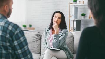 A woman sits on a couch, thoughtfully listening to two people as she holds a notepad in her lap. She appears to be a therapist, counselor, or mediator, engaged in a session with two clients. photo