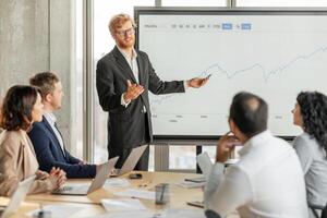 A businessman in a suit stands in front of a large screen displaying a financial chart, presenting information to his team. They are seated around a table with laptops and papers photo