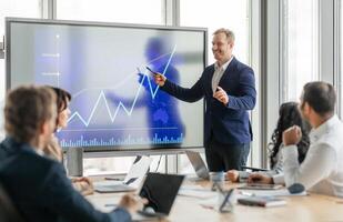 A businessman stands at the front of a meeting room, pointing at a large screen displaying a financial graph. He is presenting data to his colleagues, who are seated around a conference table. photo