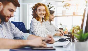 Cheerful redhead girl smiling at camera, working at coworking with other people, empty space photo