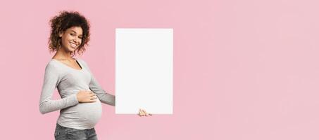 Smiling expectant woman with blank white paper touching her belly, pink background, copy space photo