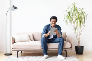 African American man is seated on a couch engaging with a tablet device in this indoor setting, showcasing modern technology and leisure in a home environment. photo