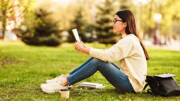 Asian woman student is seated on the green grass, engrossed in reading a book. She is alone, focused on the pages in front of her, with a serene expression on her face. photo