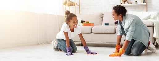 Two pretty african girls cleaning carpet at home, helping parents to keep house tidy, panorama with copy space photo