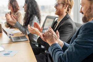 A group of business professionals are seated around a conference table, clapping their hands in appreciation. A laptop and papers are visible on the table. photo