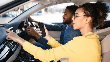 A young black woman is sitting in the passenger seat of a car. She is using her phone, likely a navigation app, as she points to the screen and smiles. The driver, a man, is looking straight ahead photo