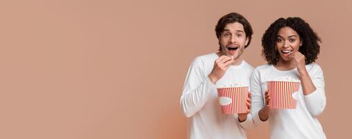 Young multiracial couple eating popcorn from buckets and looking at camera, ready to watch movie, yellow background with empty space photo