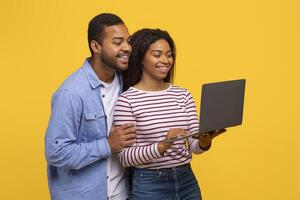 Black man and woman standing side by side, both looking intently at a laptop screen in front of them. They appear focused and engaged in whatever content is being displayed on the computer. photo
