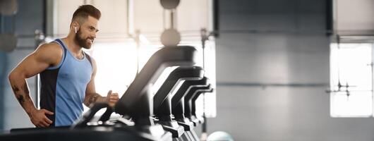 Jogging with pleasure. Young handsome man in modern wireless headphones smiling, running on treadmill at gym, free space photo