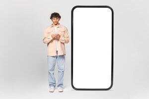 A young man in casual clothing stands in front of a large blank screen, using his smartphone. The screen is large and blank, allowing for space to add your own text or graphics. photo