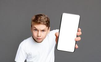 A young boy with a serious expression holds out a smartphone with a blank screen towards the camera on a gray background photo