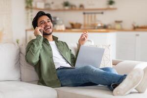 A young Arab man wearing a green button-down shirt and jeans is sitting on a couch, talking on a phone while holding a laptop. He is smiling and looking up. He is relaxed and enjoying his time. photo