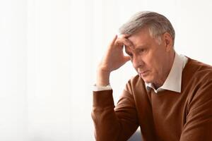 Stress. Depressed Senior Man Thinking About Problems Touching Head Sitting Against Window At Home. Selective Focus, Empty Space photo