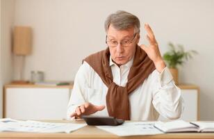 Angry Senior Man Calculating Bills And Taxes Looking At Calculator Gesturing With Hands Sitting At Work Desk Indoor. Selective Focus photo