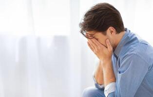 A man sits in front of a window, head bowed and hands covering his face, expressing sadness or despair. photo