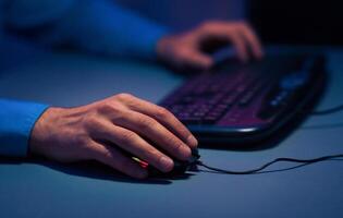 A close-up image of a persons hand using a computer mouse. The hand is in focus, while the keyboard and the persons other hand are out of focus. The lighting is dim, suggesting it is nighttime. photo