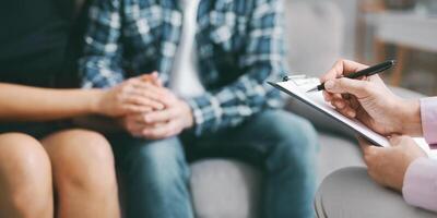 The image shows a therapist taking notes on a clipboard with a pen, while a couple sits together in a counseling session. They are holding hands, suggesting a close connection and shared experience photo