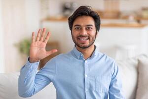 Arab man in a light blue shirt smiles and waves hello with his right hand, standing indoors in a home setting with a white couch partially visible in the background. photo