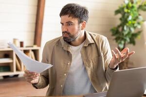 A man with a beard exhibits a puzzled expression as he examines papers in home office, laptop is open in front of him, he is cross-referencing information or dealing with an unexpected issue. photo