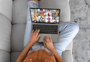 Unrecognizable lady is sitting on a couch and using a laptop to call with several other people. The person is using the trackpad on the laptop to navigate the call. photo
