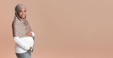 Expecting baby. Portrait of pregnant afro muslim woman in headscarf embracing her belly, posing over yellow background in studio, copy space photo