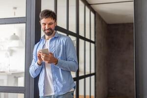 A man in a blue shirt is standing in an office hallway, leaning against a wall. He is looking down at his phone and smiling, copy space photo