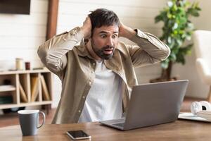 A man is seated at a wood table, hands on his head with an expression of shock and disbelief as he looks at his laptop screen. photo