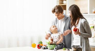 A family is cooking together in the kitchen. The father is holding a baby who is stirring a bowl of salad with a wooden spoon. The mother is standing next to them, holding a tomato, copy space photo