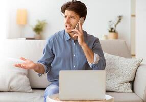 A man sits on a couch with a laptop in front of him, gesturing with one hand while talking on the phone with a frustrated expression. photo