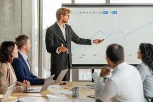 A businessman, wearing a suit, stands in front of a large screen displaying a financial chart, gesturing towards the data. He is presenting to a group of colleagues, who are seated at a table photo