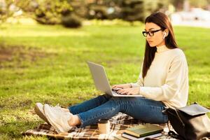 Asian woman is seated on a blanket outdoors, typing on a laptop. She is focused on the screen, surrounded by a open space. The sun is shining, and there are trees in the background. photo