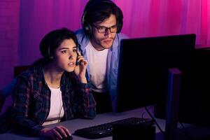 A young couple, both wearing headphones, sit in front of a computer screen in a dimly lit room. The man is standing behind the woman, both looking intently at the screen, likely engaged in a game. photo