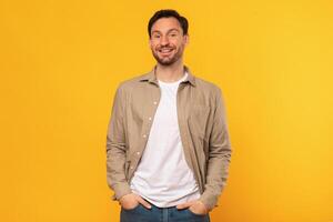 A man wearing a brown button-up shirt and jeans stands against a yellow background. He is smiling directly at the camera with his hands in his pockets. photo