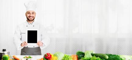 Cooking App. Chef Man Showing Digital Tablet With Empty Screen Advertising Menu Or Restaurant Application Standing In Kitchen Indoor. Mockup photo