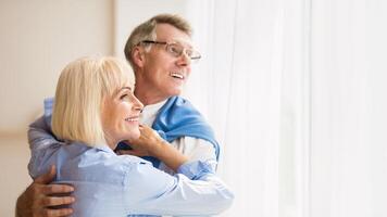 Happy senior couple looking through window and hugging at home, free space photo