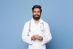 A cheerful Indian doctor in a white lab coat and stethoscope stands against a blue background, smiling as he interacts with his smartphone photo