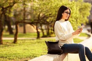 Asian woman is seated on a bench, engrossed in her cell phone. She appears focused on the screen, with her fingers tapping away as she texts or scrolls through content. photo