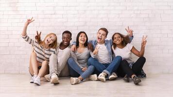 Emotional multiracial students sitting on floor, showing peace gesture, posing at camera, white wall background photo