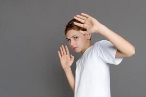 In this image, a teen boy adopts a serious expression and makes a stop gesture with his hands, signaling a boundary or refusal, isolated on a grey background photo
