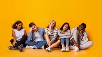 Bored international classmates sitting over yellow studio background, being tired after classes, panorama photo