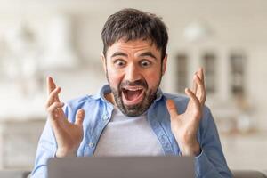 A man with a surprised expression sits in front of a laptop, his hands raised in excitement as he looks at the screen. photo