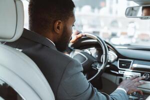 A man in a suit is driving a car, his hand resting on the dashboard. The man is focused on the road ahead, his expression serious. The city can be seen in the background photo