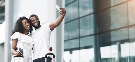 African American couple taking a selfie at an airport terminal. The man is holding his smartphone, and the woman is smiling at the camera while holding a passport and boarding pass, copy space photo